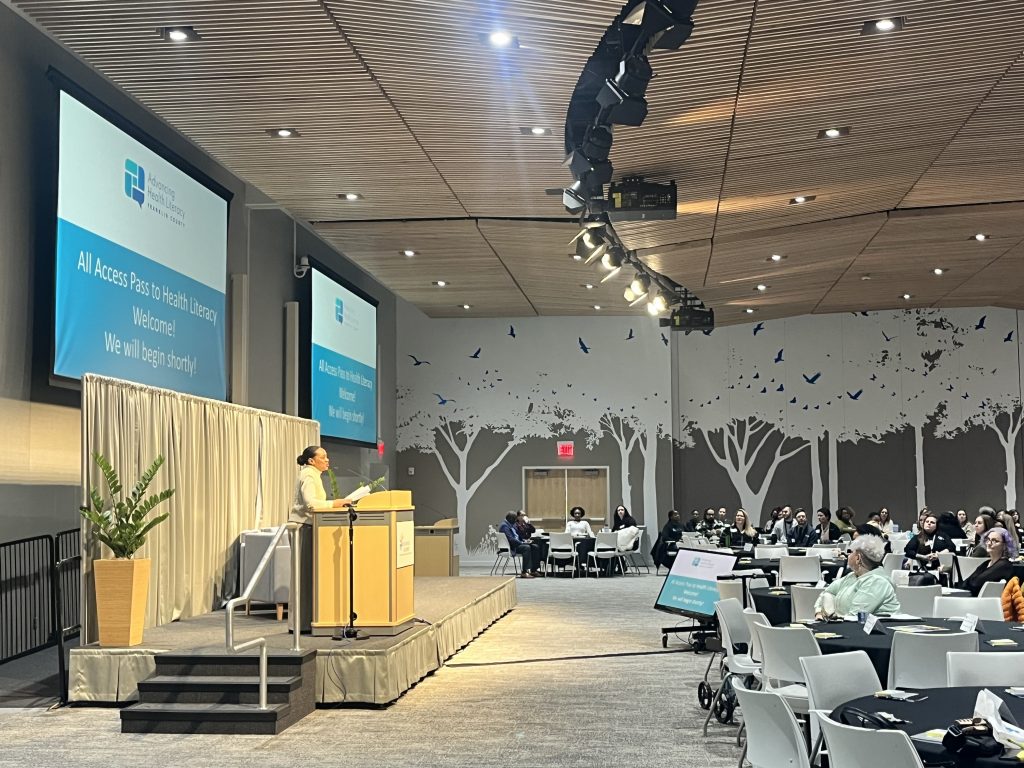 Photo of a woman speaking at a podium to an audience of people